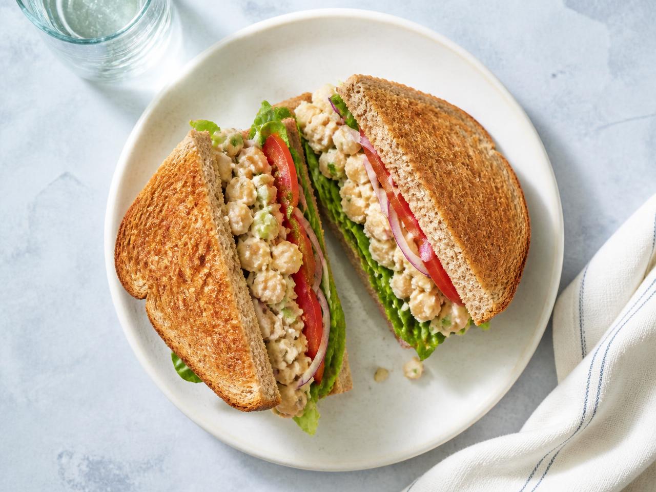 Chickpea no-tuna salad sandwich on toasted whole wheat bread with lettuce, tomato, and red onion, cut in half on a white plate